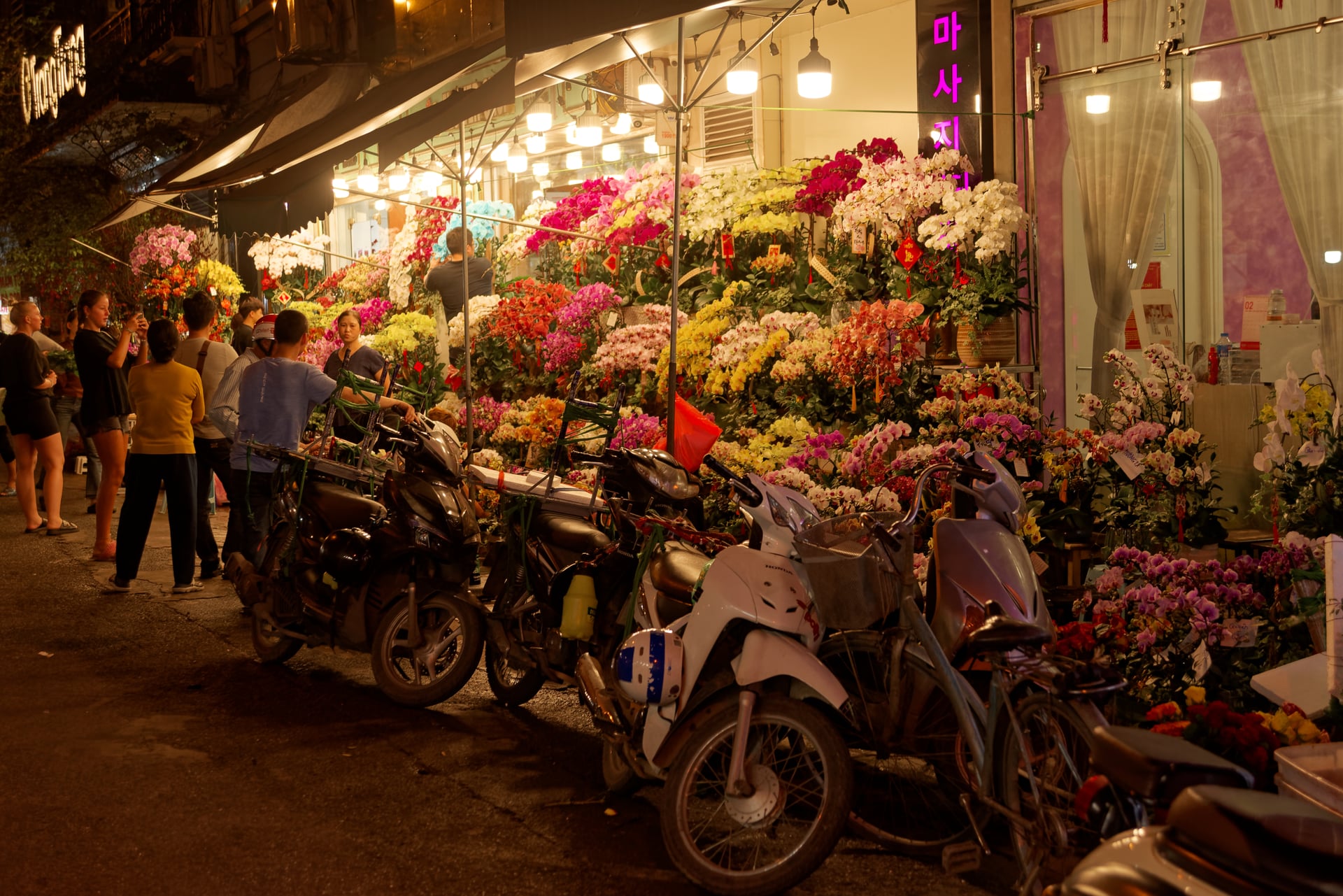 河內老城夜裡的花攤與行人,暖色吊燈照亮街道,前景停放機車